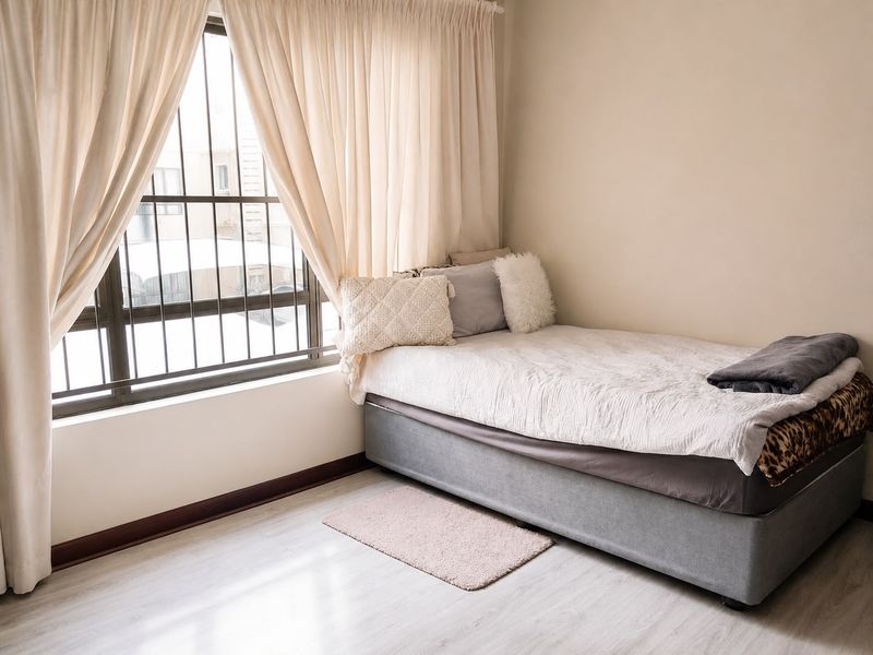 A bright bedroom featuring light-toned laminate flooring, neutral walls, and a large window fitted with security bars.