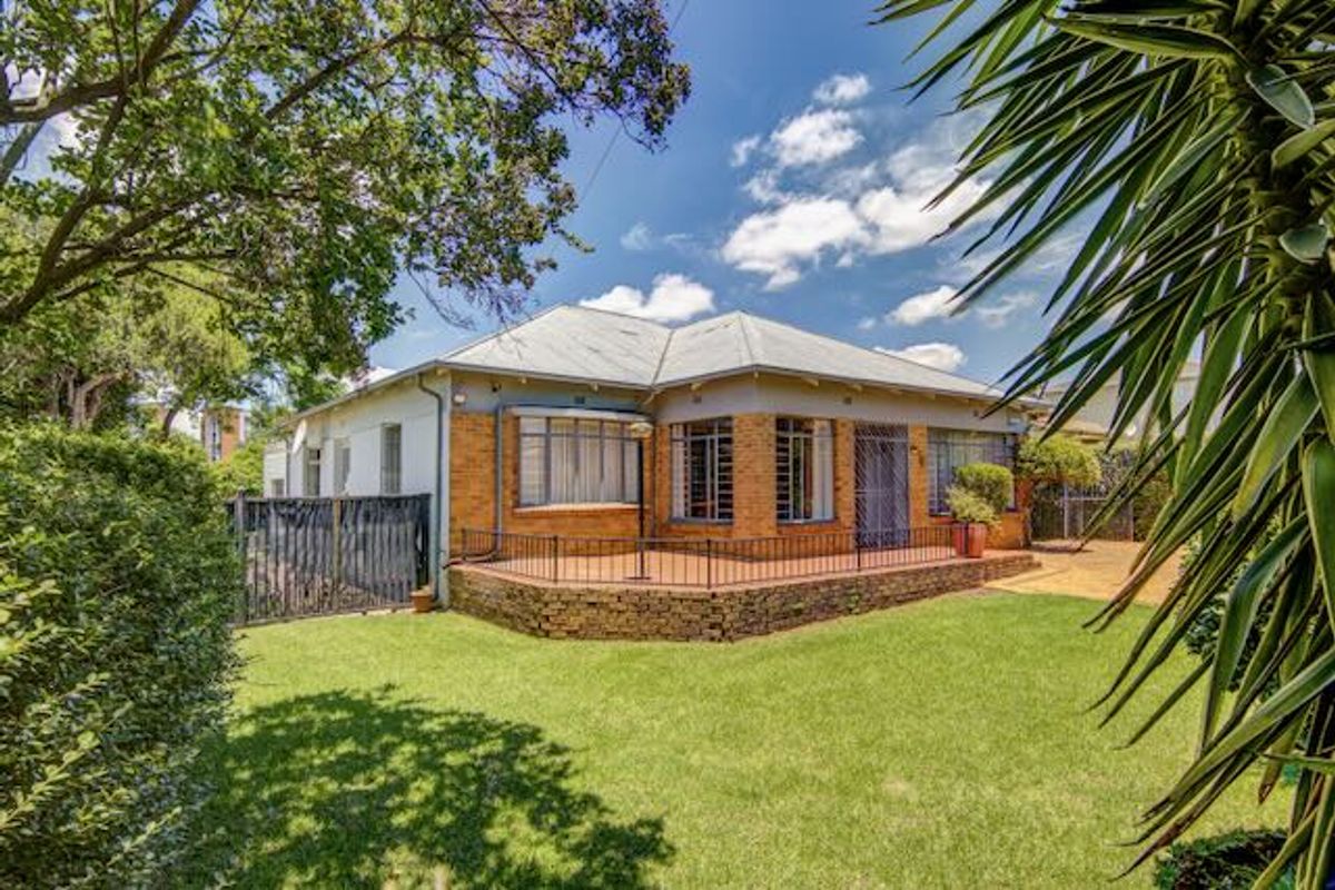 A well-maintained brick house featuring a raised stone patio with wrought iron railings, overlooking a spacious green lawn and mature garden.