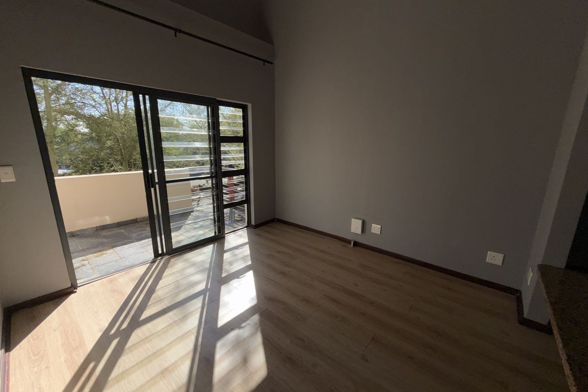 A bright, empty living area featuring modern laminate flooring, neutral grey walls, and a large sliding glass door providing access to an outdoor balcony.