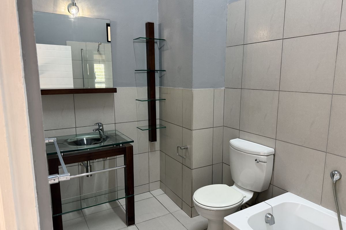 A clean, modern bathroom featuring a glass vanity basin, wall-mounted mirror, toilet, and bathtub with neutral wall and floor tiling.