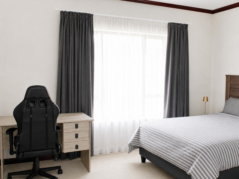 A well-maintained bedroom featuring neutral carpet flooring, timber crown molding, and large windows dressed with floor-to-ceiling curtains.