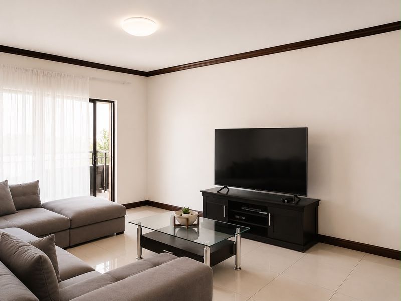 A bright and well-maintained living room featuring neutral tiled flooring, dark crown molding, and a sliding glass door providing access to the balcony.