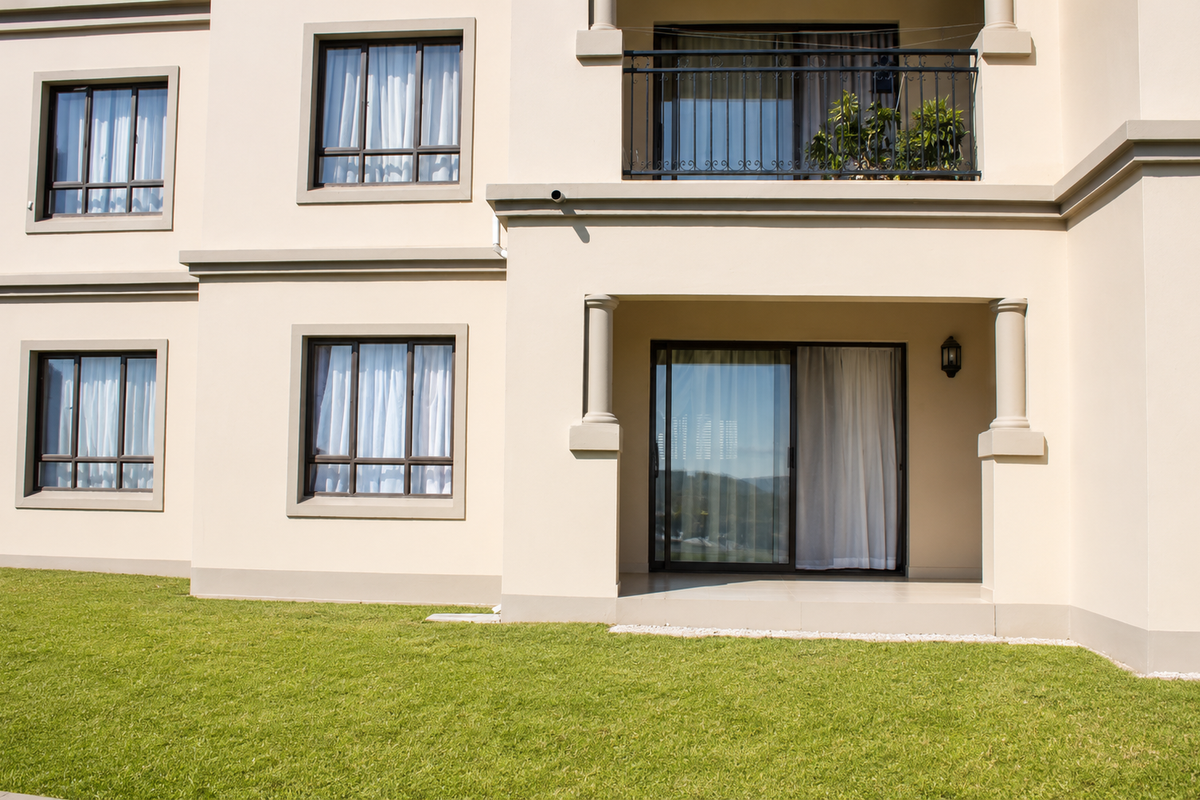 Rear elevation of the apartment building featuring a ground floor patio, upper balcony with wrought iron railing, and well-maintained lawn area.