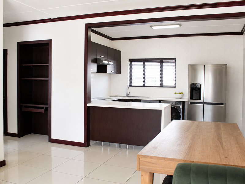 Open-plan kitchen area featuring dark wood cabinetry, white countertops, tiled flooring, and integrated storage shelving, seamlessly connecting to the dining space.