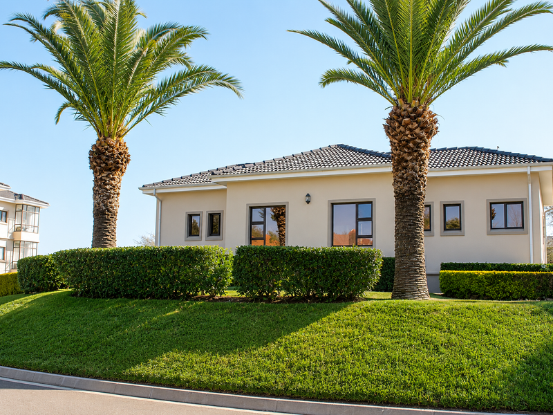 A well-maintained single-story apartment exterior featuring a tiled roof, modern aluminium window frames, and manicured garden landscaping with mature palm trees.