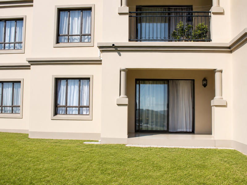 Rear elevation of the apartment building featuring a ground floor patio, upper balcony with wrought iron railing, and well-maintained lawn area.