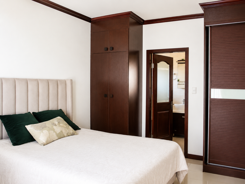 A well-maintained bedroom featuring dark wood built-in cabinetry, crown molding, and direct access to an en-suite bathroom with tiled flooring throughout.