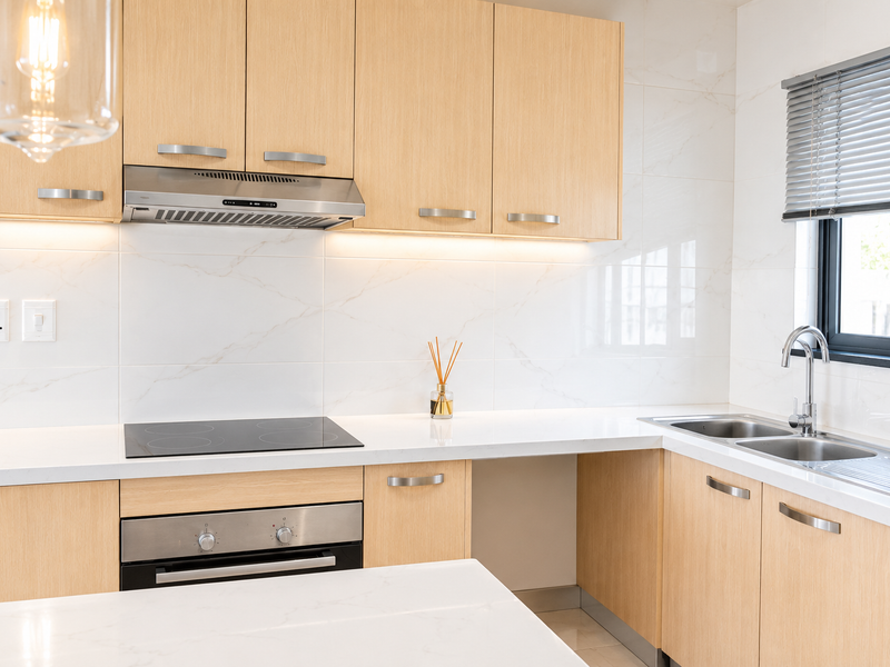 Modern kitchen featuring light-wood cabinetry, white marble-look backsplash, quartz countertops, integrated stainless steel appliances, and a double sink with a window view.