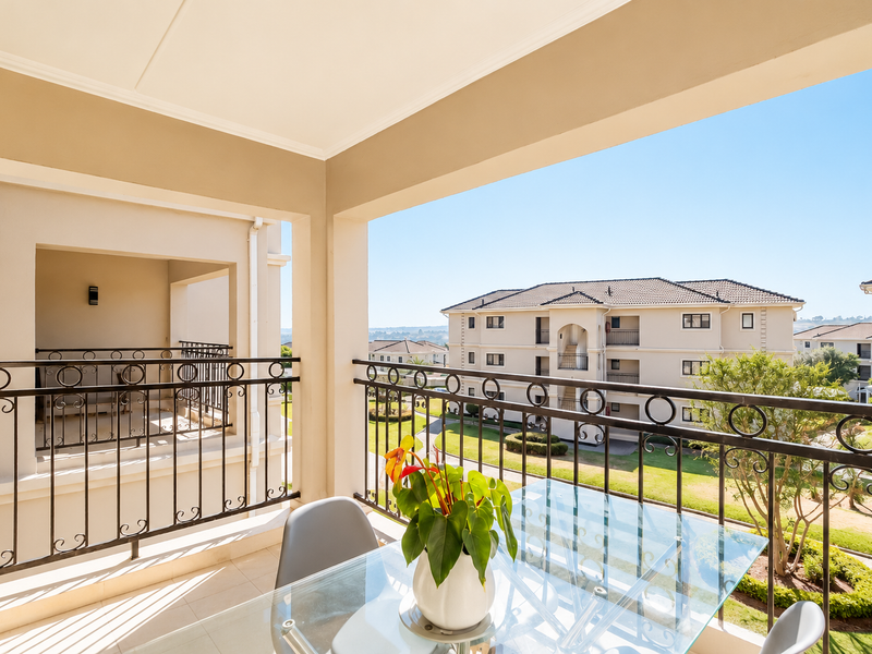 A spacious covered balcony featuring a decorative wrought iron balustrade and tiled flooring, offering pleasant views of the surrounding apartment complex grounds.