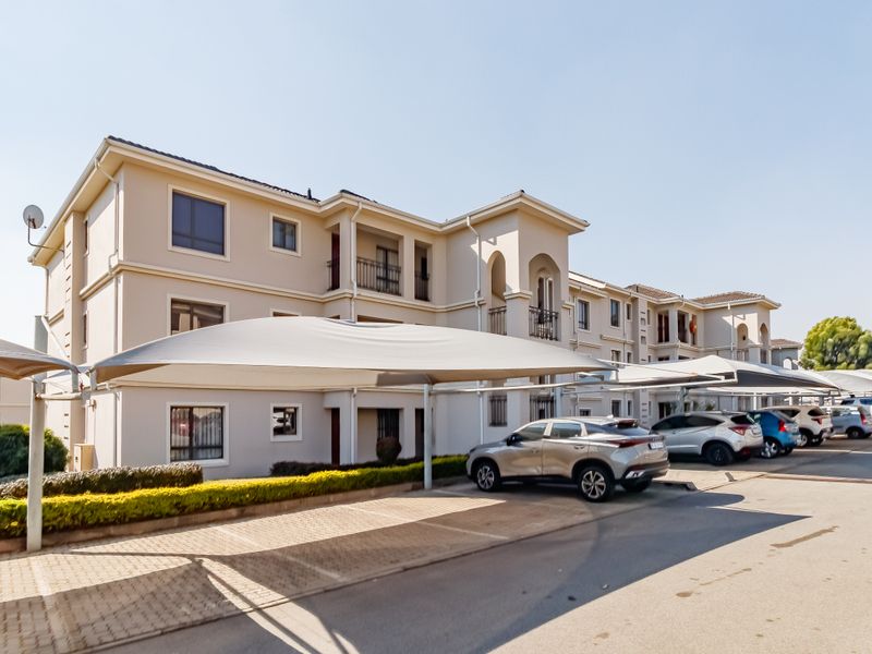 Exterior view of a multi-story apartment complex featuring paved parking areas with shade cloth carports and well-maintained landscaped hedging.