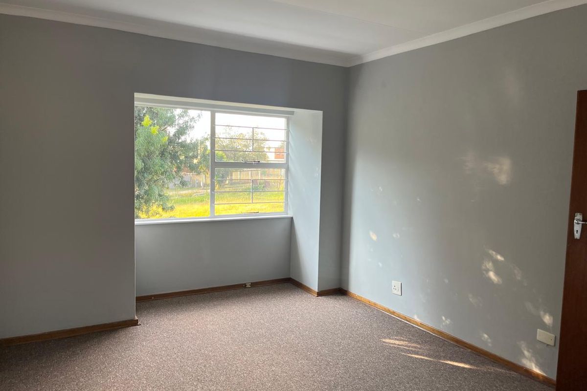 A clean, empty bedroom featuring neutral grey walls, carpeted flooring, and a window fitted with security bars.