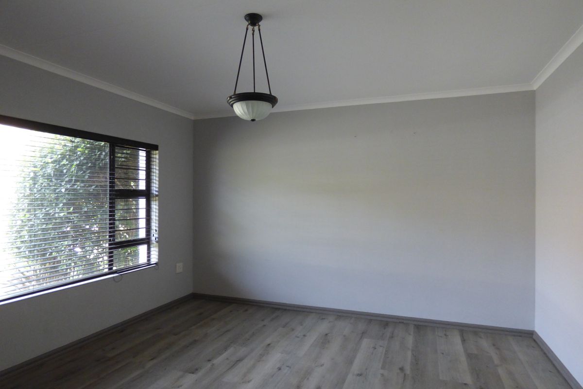 A bright, empty bedroom featuring modern laminate flooring, neutral grey walls, and a classic pendant light fixture with venetian blinds.