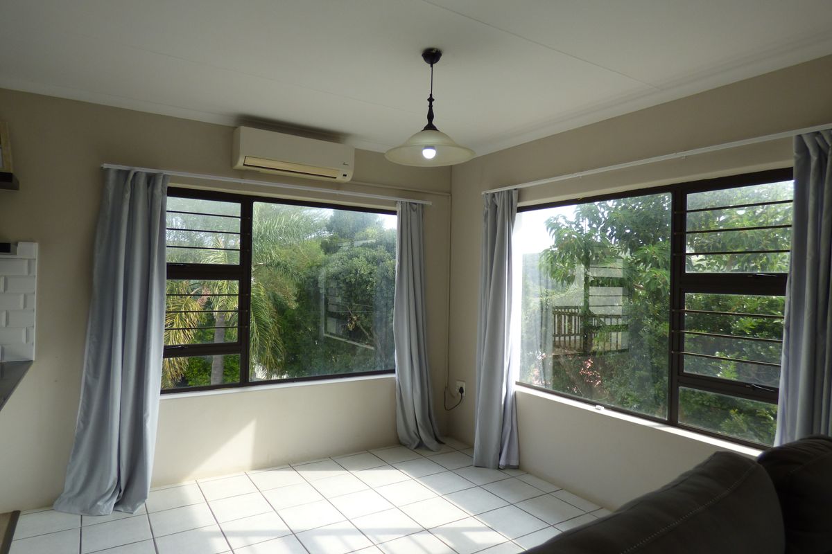 A bright living area featuring tiled flooring, large windows with security bars, and a wall-mounted split-system air conditioner.
