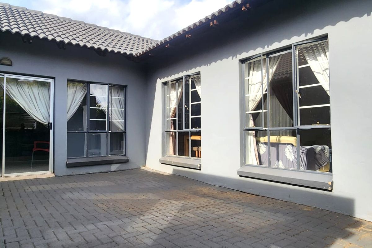A rear exterior view of the house featuring a paved patio area, grey painted walls, tiled roofing, and multiple aluminium-framed windows and a sliding door.