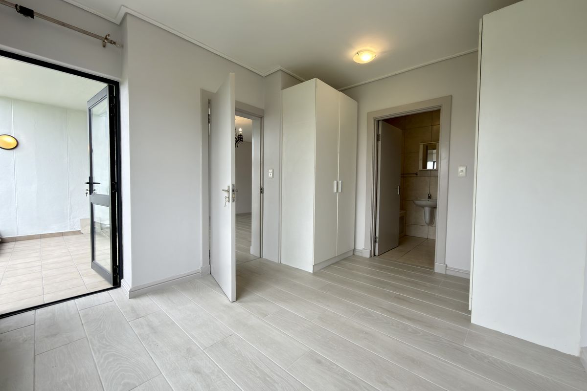 A bright bedroom featuring light wood-look tile flooring, built-in cupboards, and direct access to a private balcony, with an en-suite bathroom attached.