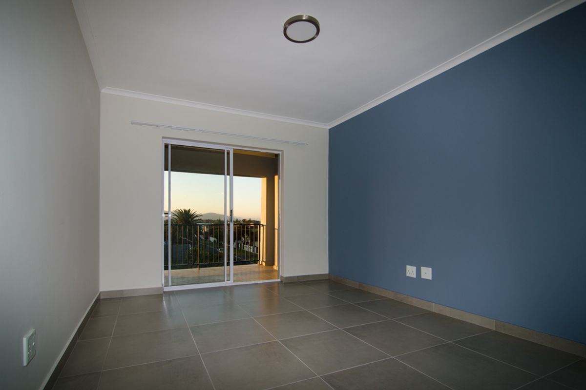 A bright, empty bedroom featuring neutral and blue accent walls, tiled flooring, and sliding glass doors leading to a private balcony.