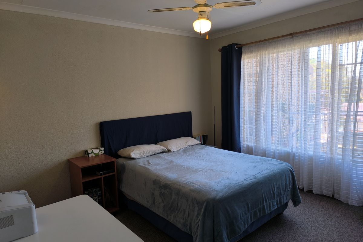 A functional bedroom featuring a ceiling fan, carpeted flooring, and large windows with curtain rods, providing a comfortable and well-lit living space.