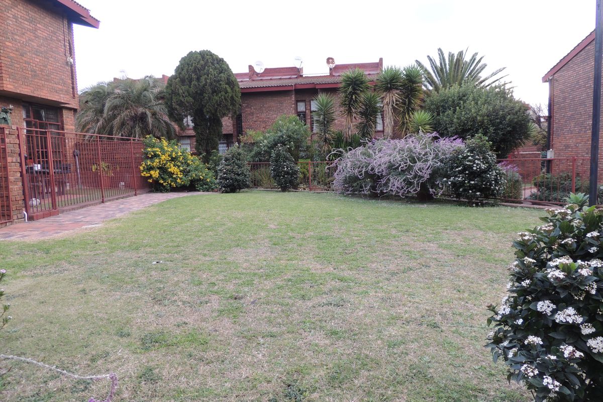 A view of the rear garden area featuring a well-maintained lawn, established shrubbery, and brick townhouse exterior walls with metal fencing.
