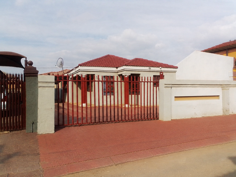 Front view of a residential property featuring a red tiled roof, secure perimeter wall with metal gate, and a paved driveway.