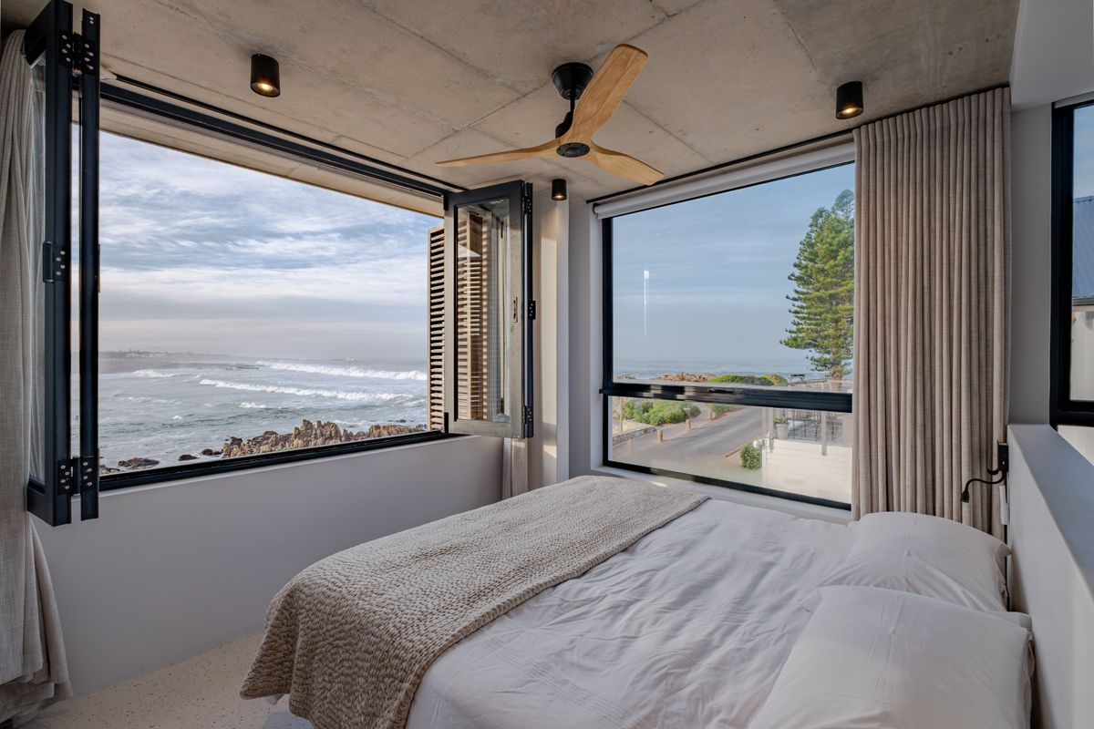 Modern bedroom featuring an exposed concrete ceiling, ceiling fan, and expansive windows offering panoramic ocean views.