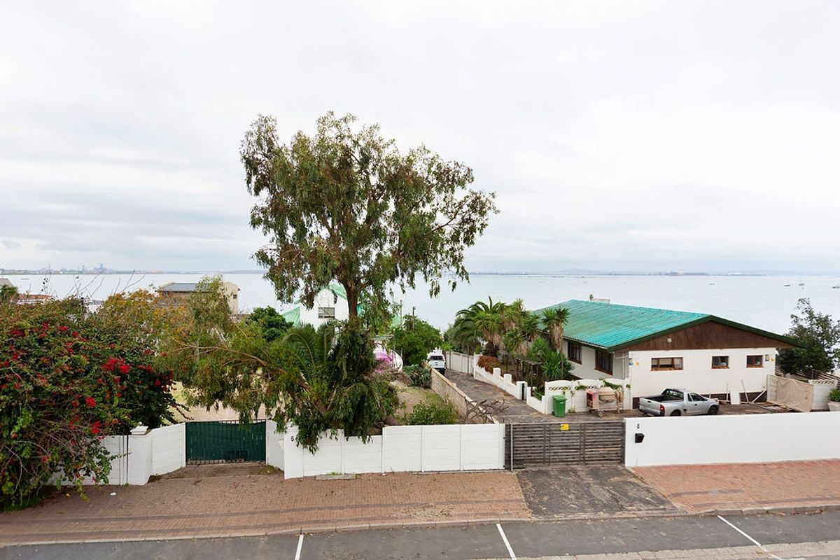 Aerial view of a coastal property featuring a paved driveway, boundary walls, and a pitched metal roof with scenic water views.