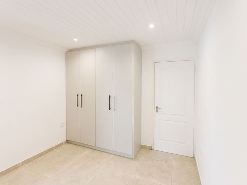A clean, bright bedroom featuring neutral tiled flooring, a modern built-in wardrobe, and a white tongue and groove ceiling with recessed lighting.