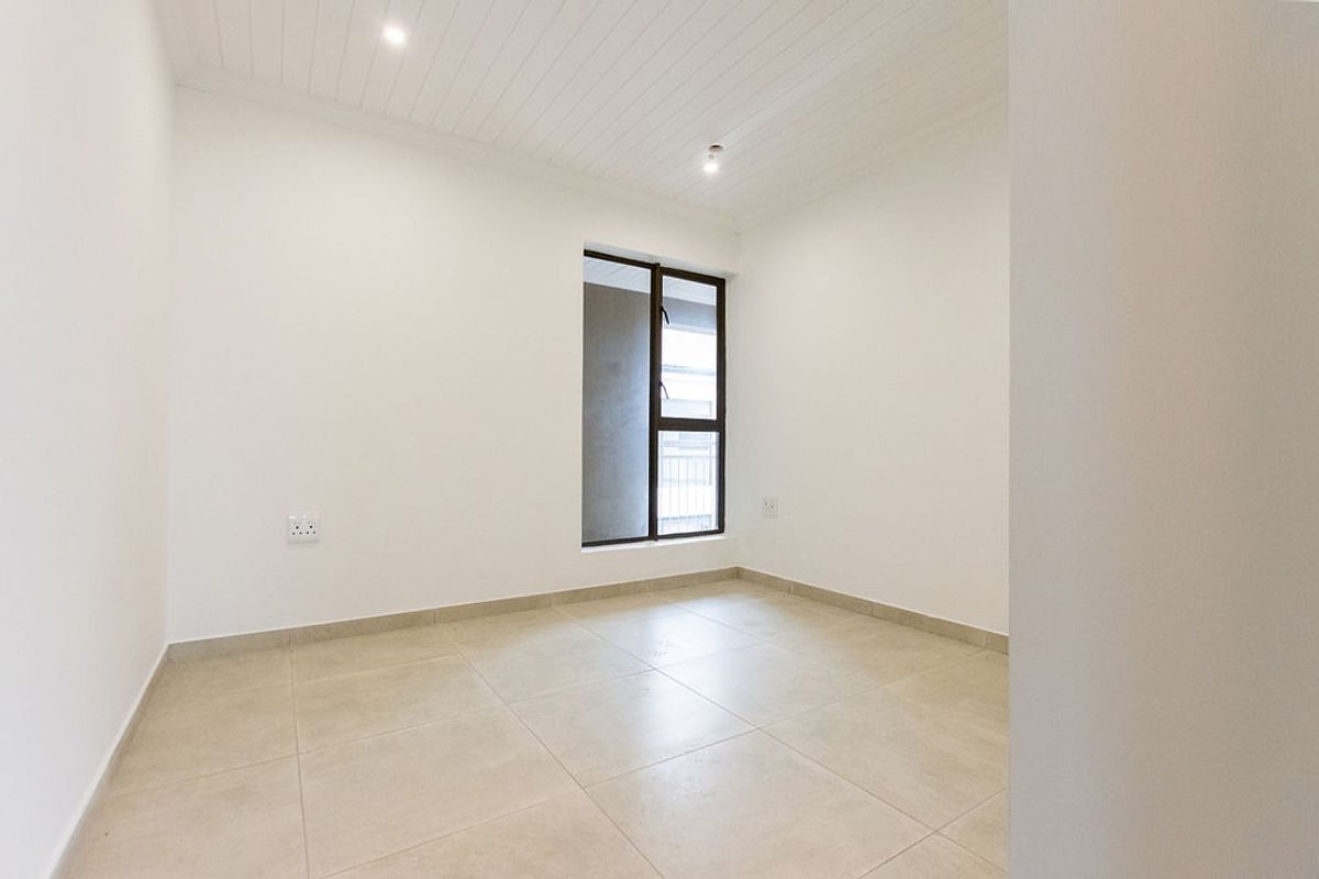 A bright, empty bedroom featuring neutral tiled flooring, a tongue and groove ceiling with recessed lighting, and a large aluminium-framed window.