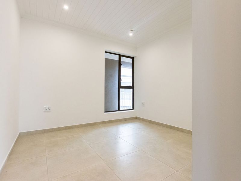 A bright, empty bedroom featuring neutral tiled flooring, a tongue and groove ceiling with recessed lighting, and a large aluminium-framed window.