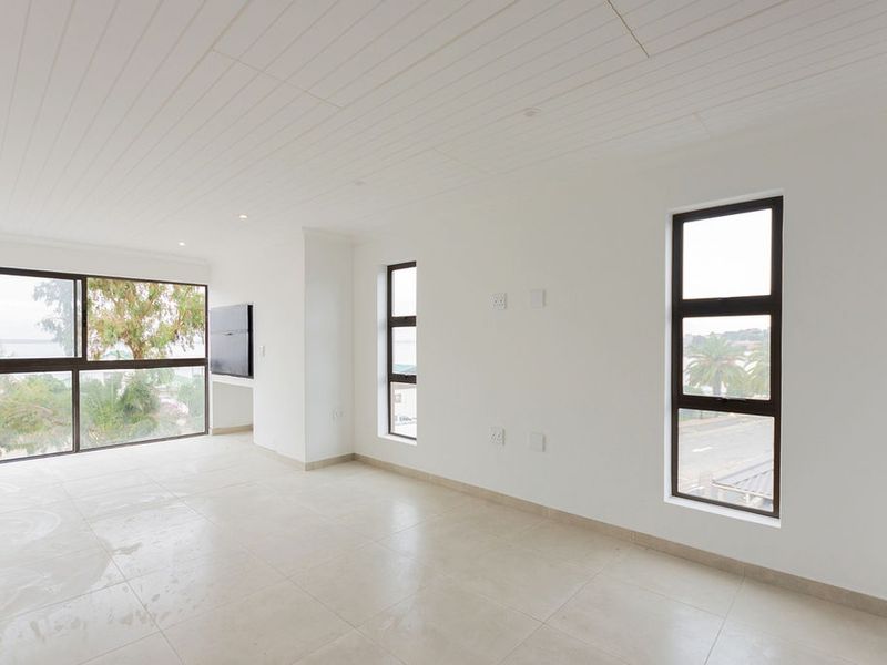 A bright, empty living area featuring tiled floors, a tongue and groove ceiling, and large aluminium-framed windows with views.