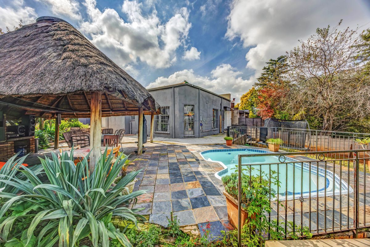 Rear garden area featuring a swimming pool with metal fencing, a slate-tiled patio, and a traditional thatch-roofed lapa for outdoor entertainment.