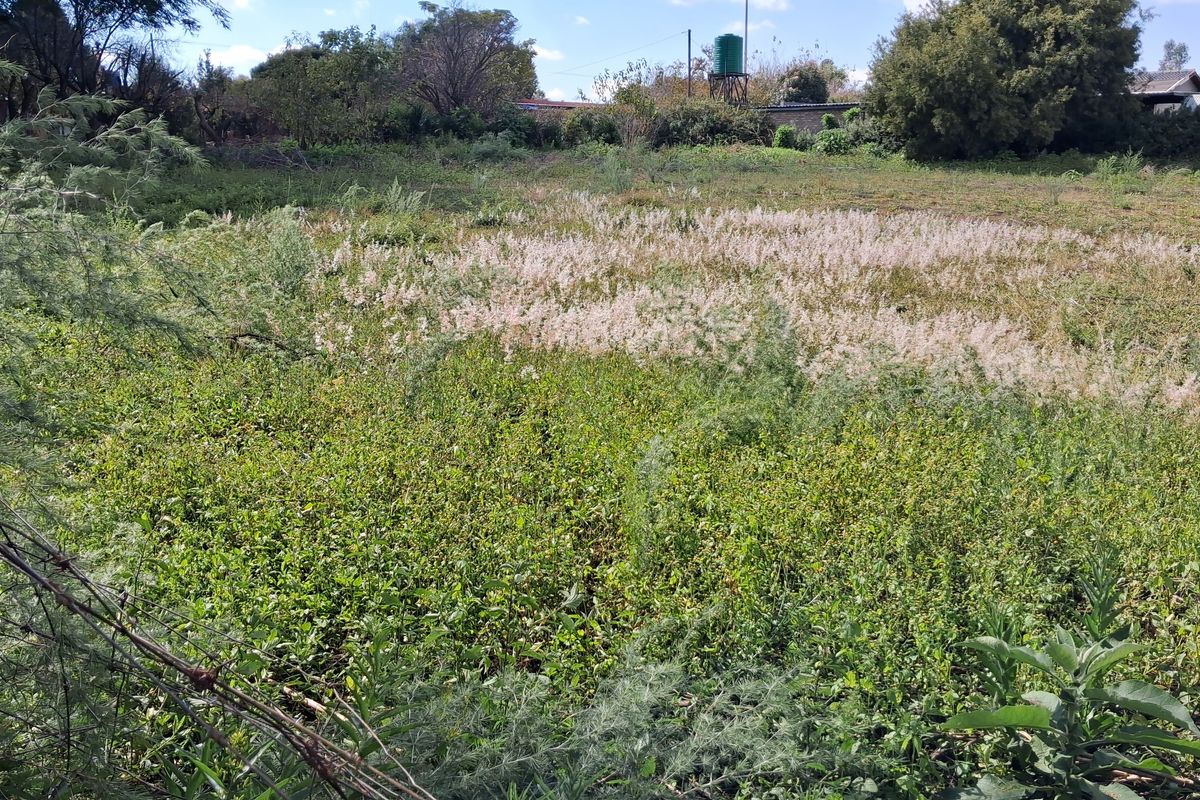 A view of a vacant residential plot featuring overgrown vegetation, perimeter fencing, and an existing water tank on the property.