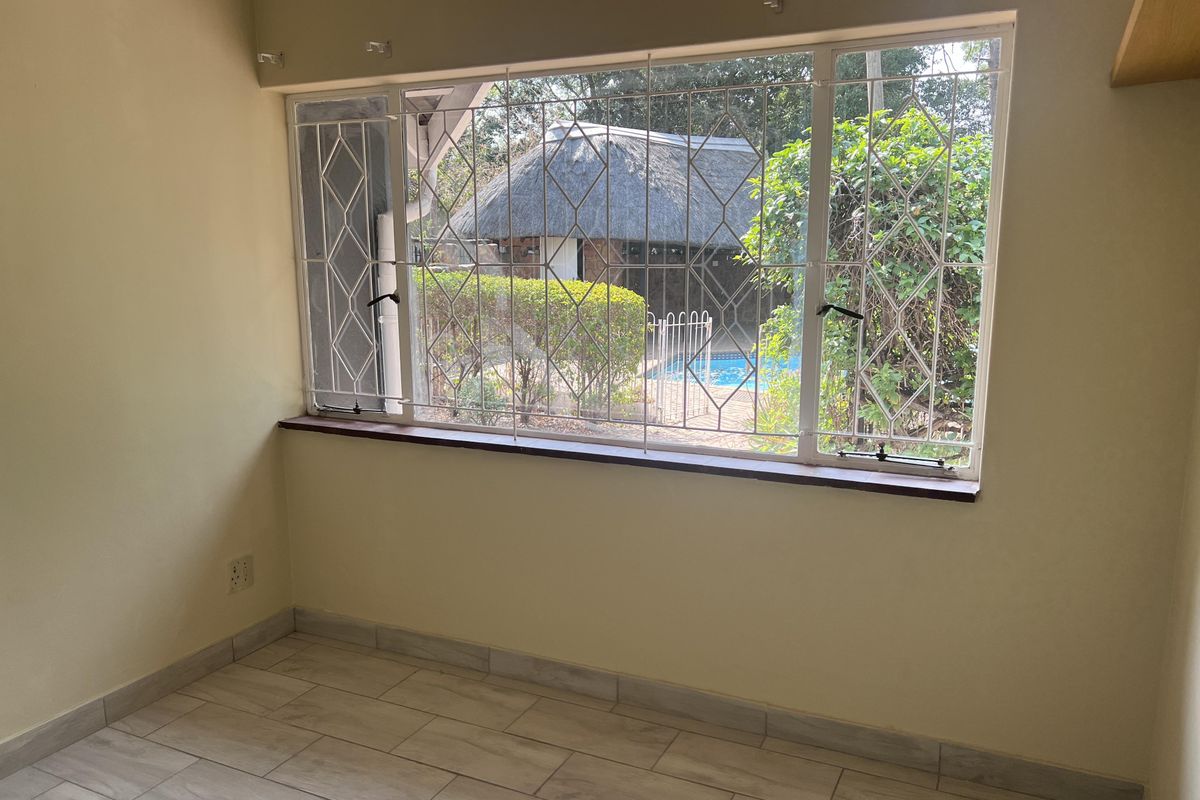 An empty bedroom featuring tiled flooring and a large window with security burglar bars, offering a view of the garden and pool area.