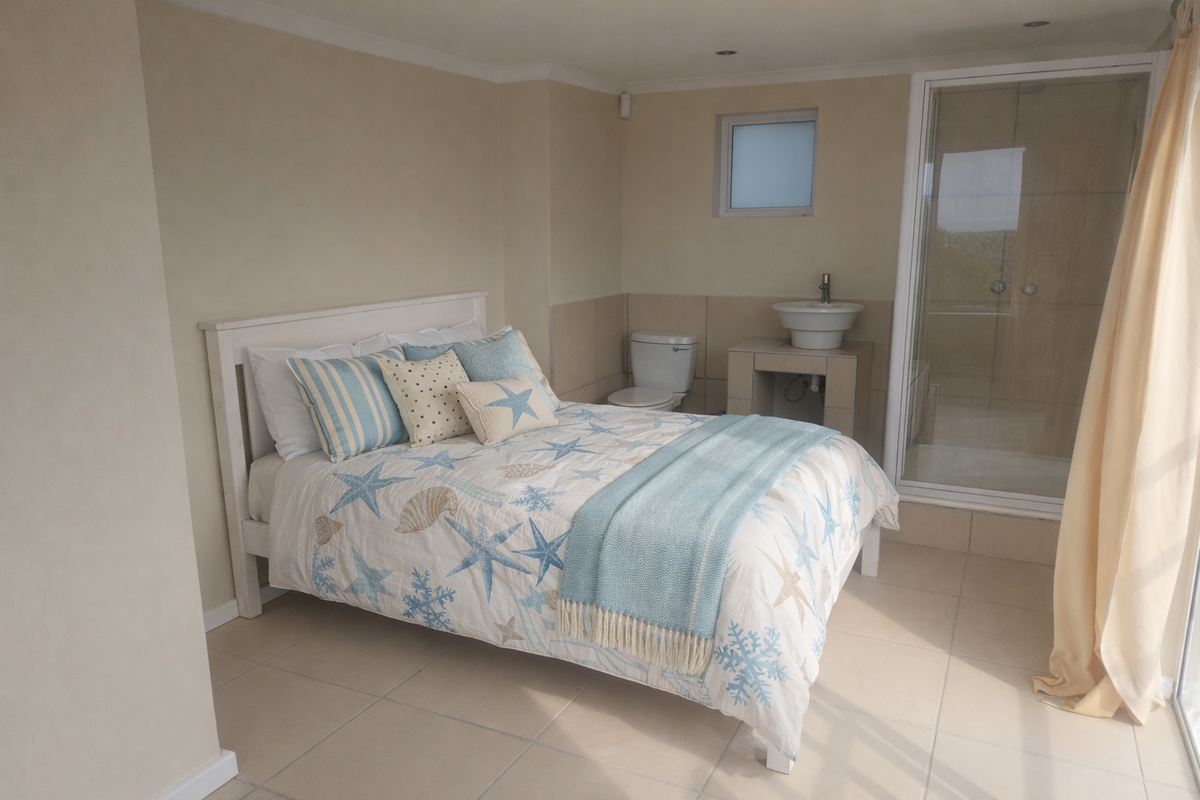A bedroom featuring an open-plan en-suite bathroom with a shower, toilet, and pedestal basin, finished with neutral wall tones and tiled flooring.