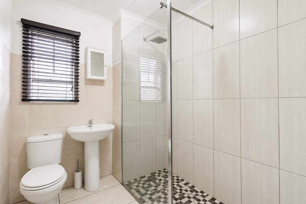 A clean, modern bathroom featuring a pedestal basin, a walk-in shower with a glass screen, and neutral wall tiling.