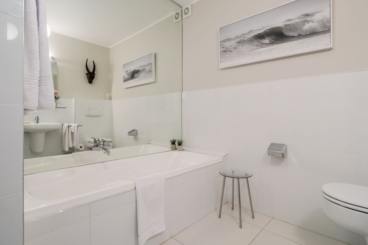 A clean, well-maintained bathroom featuring a built-in bathtub, wall-mounted toilet, pedestal basin, and large wall-to-wall mirror with white tiled finishes.