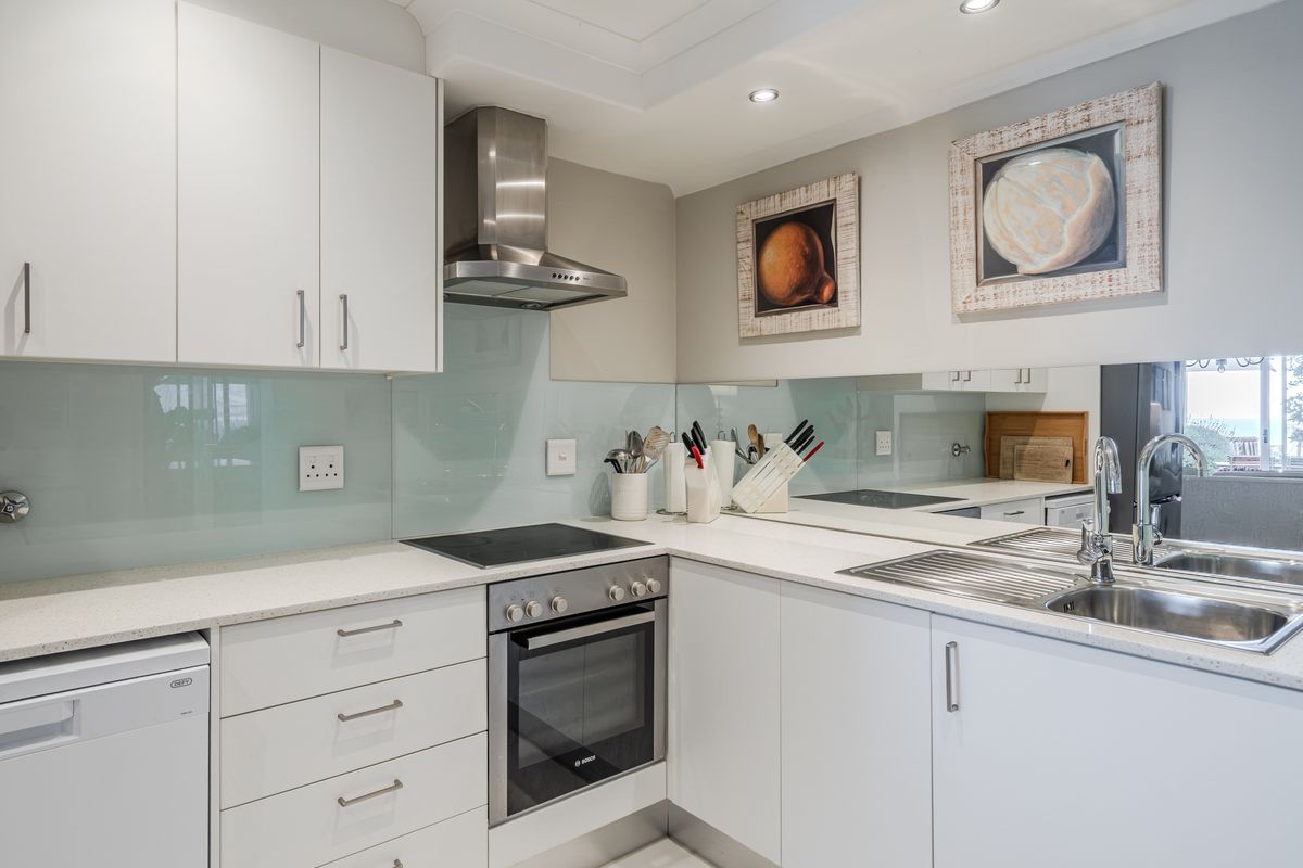 Modern kitchen featuring white cabinetry, stone countertops, a glass splashback, stainless steel extractor fan, and a built-in oven with electric hob.