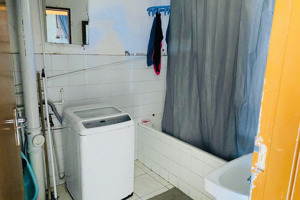 A functional bathroom featuring a bathtub, washbasin, and tiled finishes, currently utilized as a laundry area with exposed plumbing and visible wear.