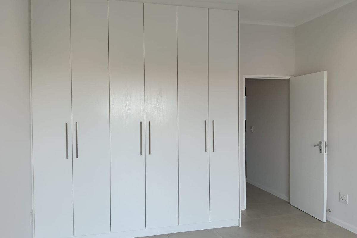 A clean, modern bedroom featuring floor-to-ceiling white built-in cupboards, neutral tiled flooring, and recessed downlighting.