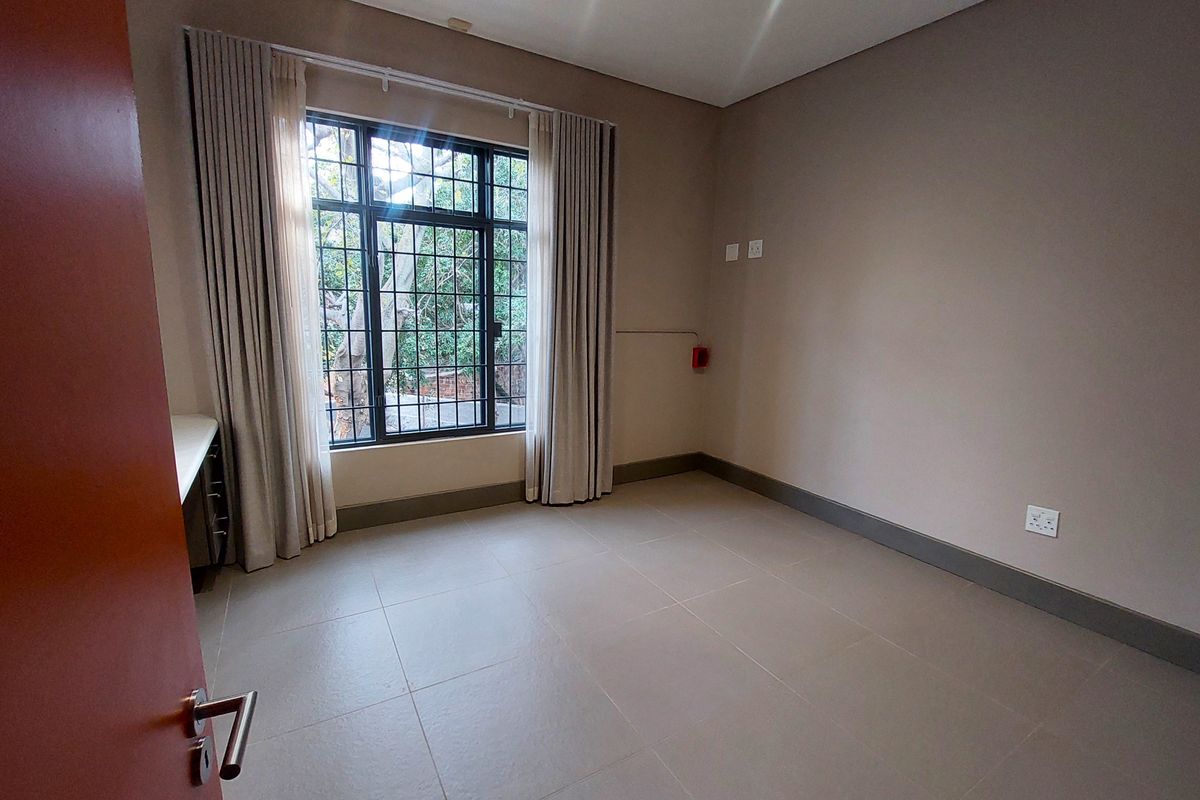 A clean, empty bedroom featuring neutral tiled flooring, a built-in desk area, and a window fitted with security bars.