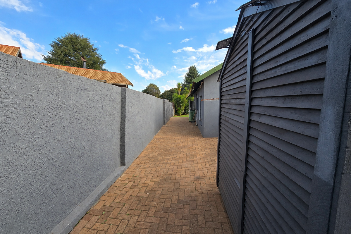 A side passage featuring a paved walkway bordered by a high boundary wall and a structure with dark timber cladding.