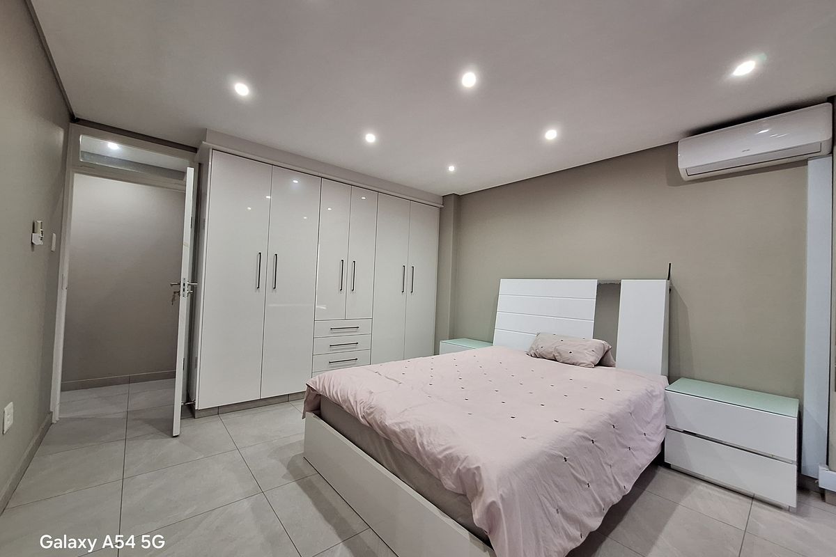 A modern bedroom featuring neutral walls, large format floor tiles, extensive built-in cabinetry, and a wall-mounted air conditioning unit.