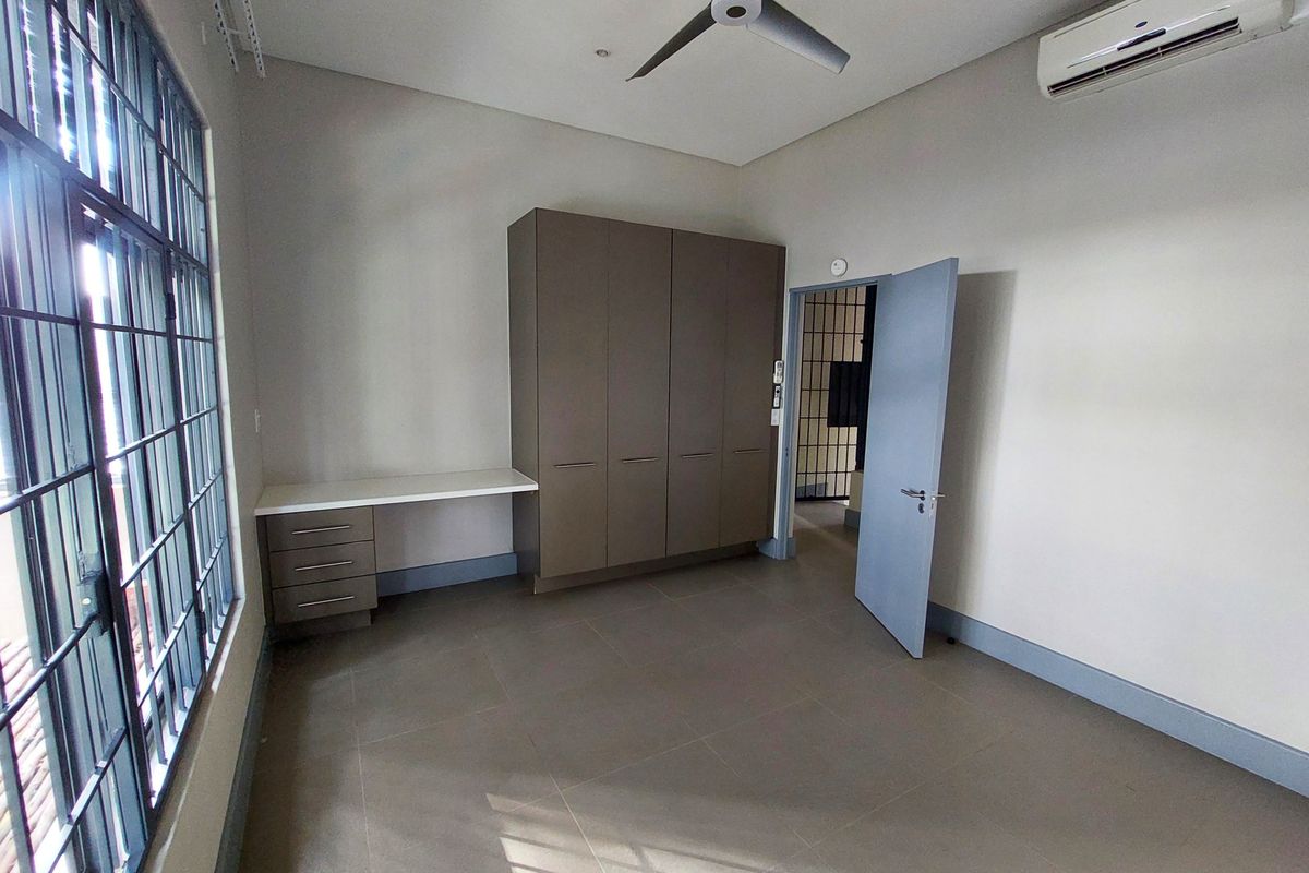 A spacious, empty bedroom featuring neutral tiled flooring, built-in cupboards with an integrated desk, a ceiling fan, and a wall-mounted air conditioning unit.