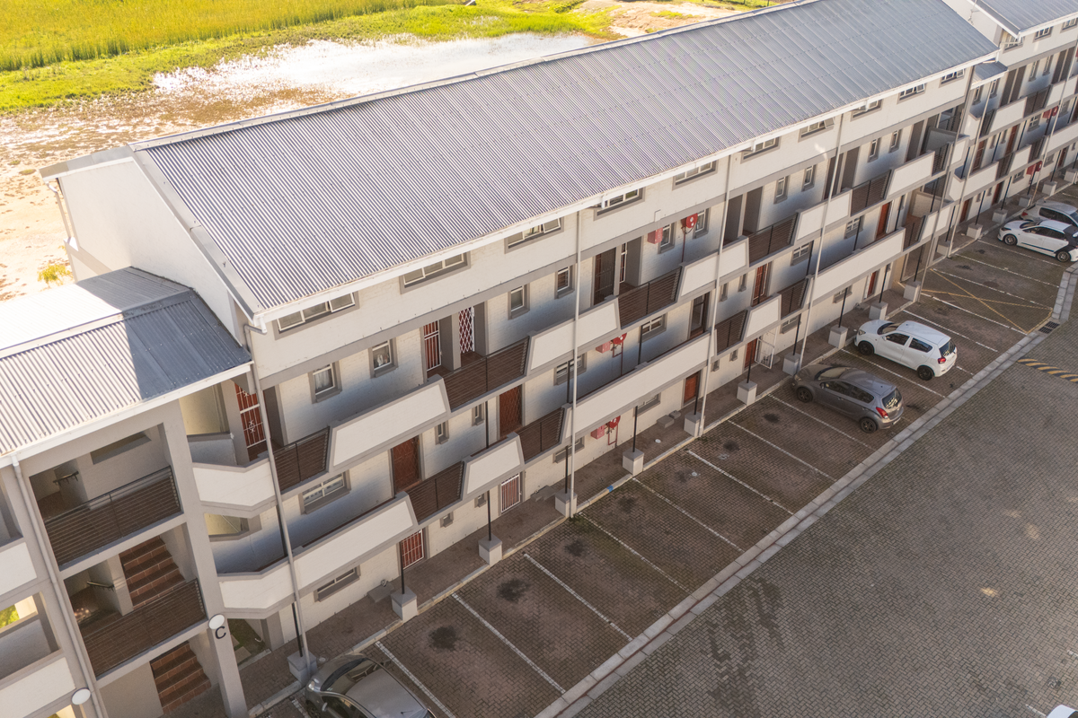 Aerial view of a multi-story apartment complex featuring a corrugated metal roof, external stairwells, private balconies, and an adjacent paved parking area.