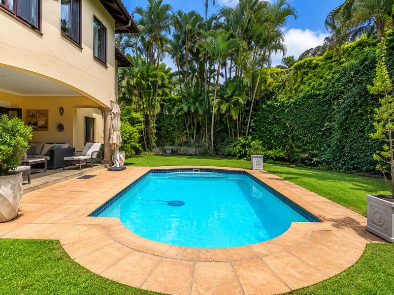 A well-maintained outdoor pool area featuring a tiled surround, covered patio space, and lush landscaped garden with mature palm trees.