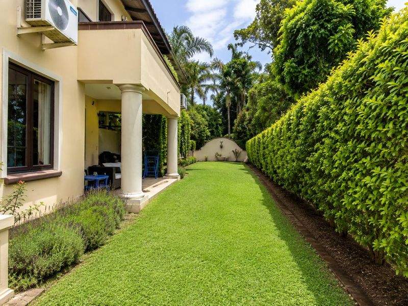 A well-maintained rear garden featuring a covered patio with classical columns, lush green lawn, manicured hedging, and a decorative wall fountain.