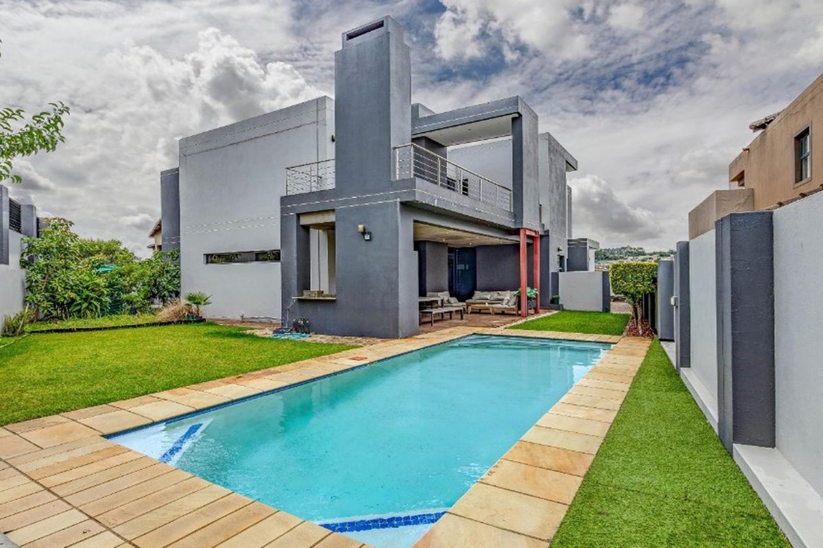 Modern rear exterior featuring a swimming pool with stone coping, artificial lawn, and a covered patio area with an integrated fireplace chimney.