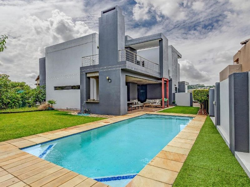 Modern rear exterior featuring a swimming pool with stone coping, artificial lawn, and a covered patio area with an integrated fireplace chimney.