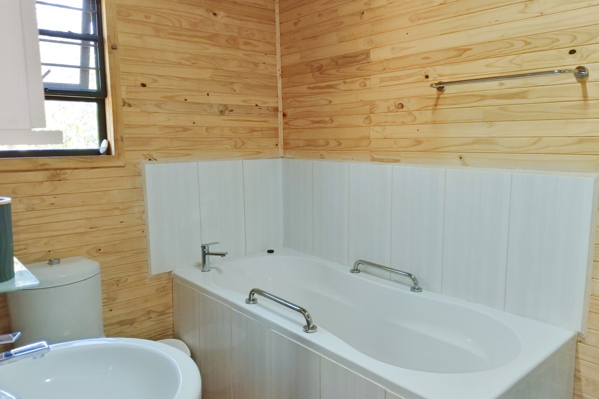 A clean bathroom featuring timber wall cladding, a white bathtub with grab rails, a pedestal washbasin, and a toilet.