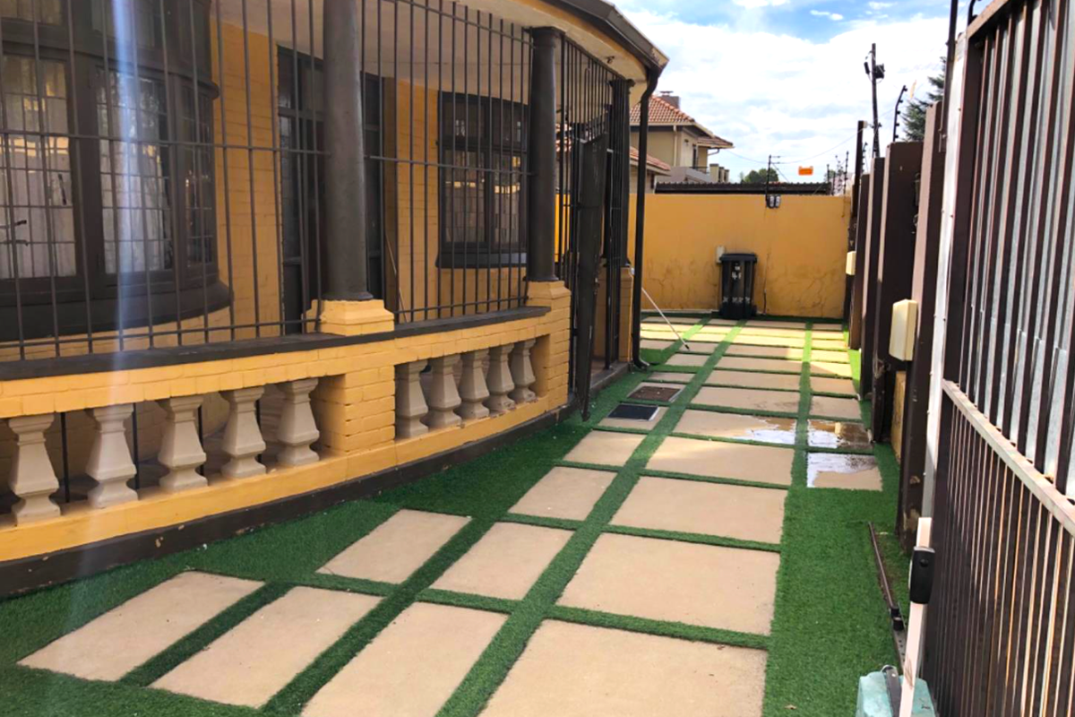 A side exterior view of the property featuring a paved walkway with artificial grass, security bars, and a yellow painted boundary wall.