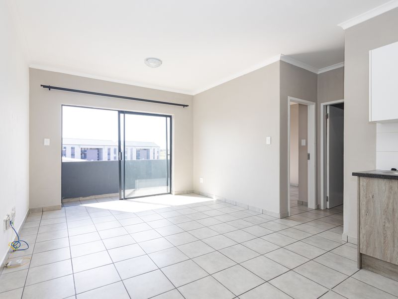 An open-plan living area featuring tiled floors, neutral walls, and a sliding door leading to a balcony, adjacent to a compact kitchen space.